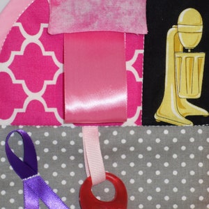 May include: A pink and gray baby activity quilt with various textures and colors. The quilt features a pink ribbon, a purple ribbon, a red textured ring, and a black and white patterned fabric with a yellow and white striped fabric.