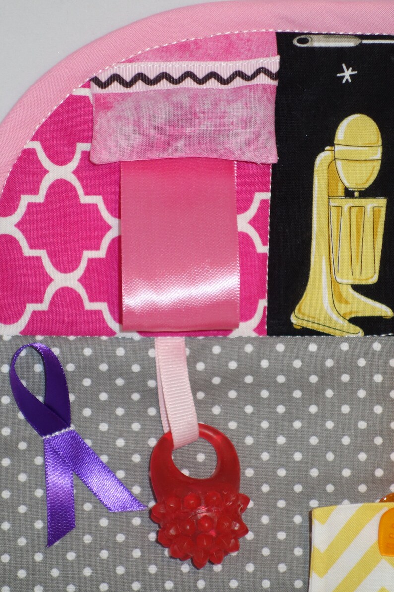 May include: A pink and gray baby activity quilt with various textures and colors. The quilt features a pink ribbon, a purple ribbon, a red textured ring, and a black and white patterned fabric with a yellow and white striped fabric.