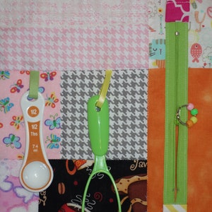 May include: A pink and white fabric activity mat with various textures and objects attached. The objects include a measuring spoon, a green potato masher, a zipper, and a string of colorful beads.