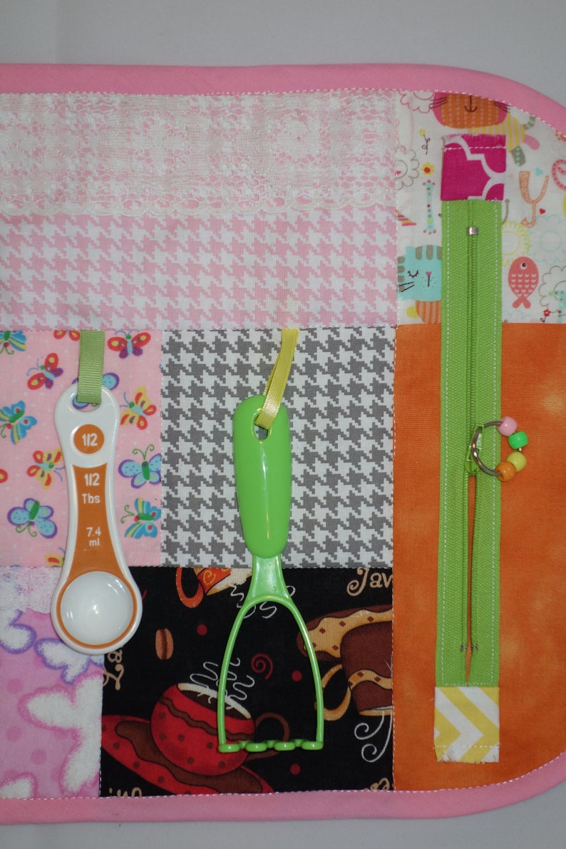 May include: A pink and white fabric activity mat with various textures and objects attached. The objects include a measuring spoon, a green potato masher, a zipper, and a string of colorful beads.
