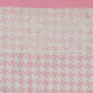 May include: A close-up of a pink and white fabric with a white lace trim. The fabric has a houndstooth pattern.
