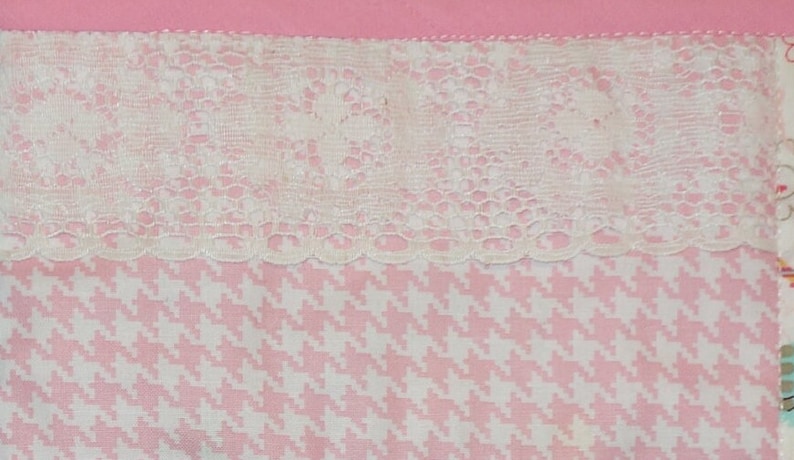 May include: A close-up of a pink and white fabric with a white lace trim. The fabric has a houndstooth pattern.