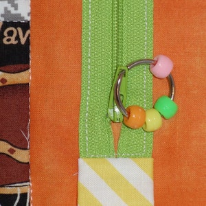 May include: A green zipper with a silver ring and colorful beads attached. The zipper is sewn into a piece of orange fabric with a yellow and white striped fabric below it.