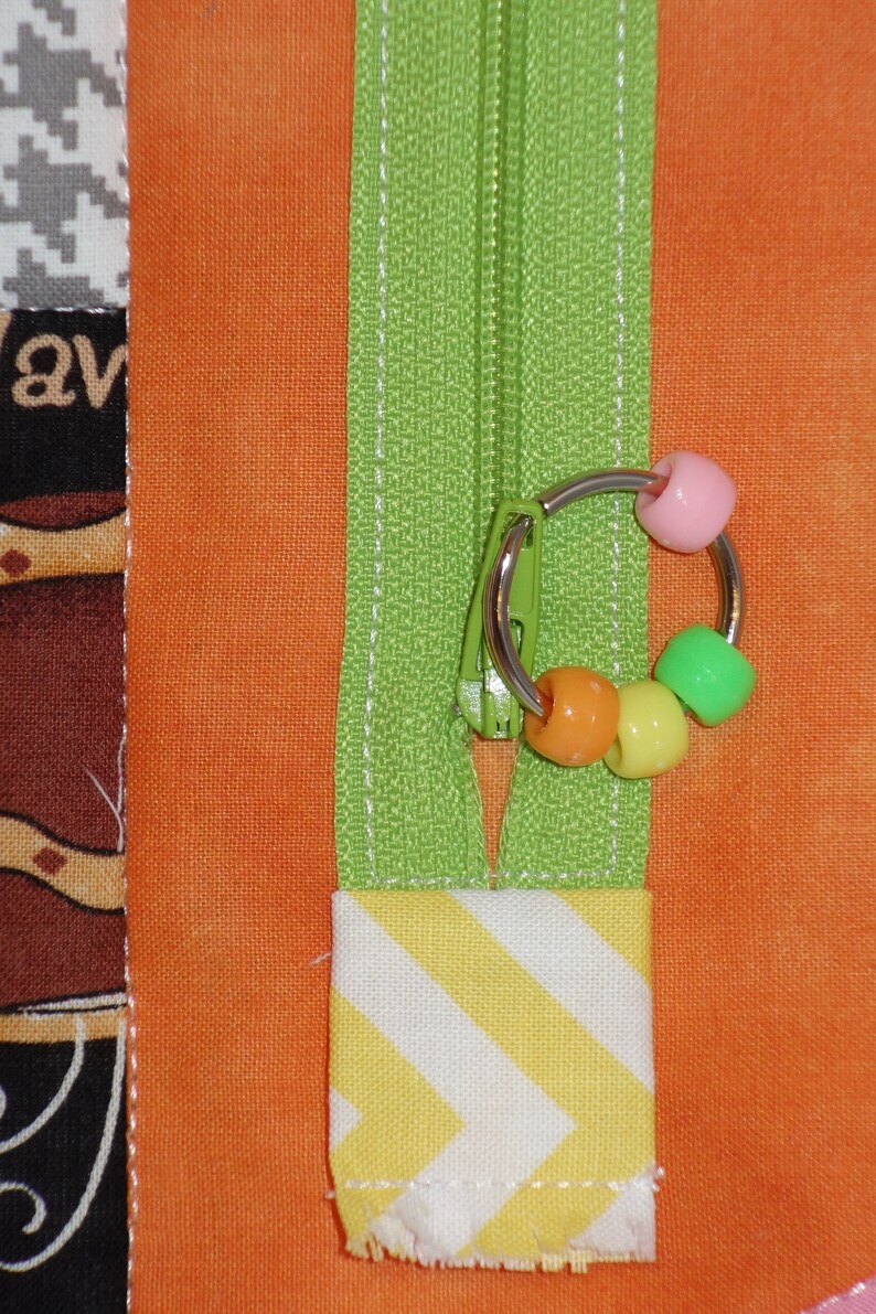 May include: A green zipper with a silver ring and colorful beads attached. The zipper is sewn into a piece of orange fabric with a yellow and white striped fabric below it.