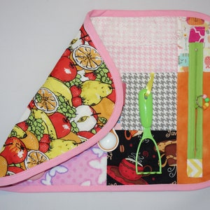 May include: A pink and white fabric activity mat with various textures and features for babies and toddlers. The mat features a zipper, a green plastic utensil, and a variety of colorful fabrics.