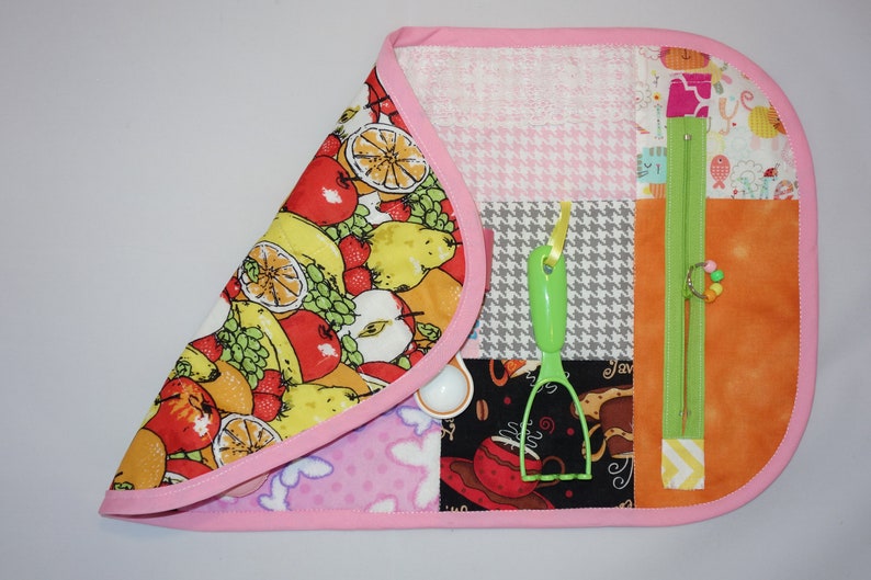 May include: A pink and white fabric activity mat with various textures and features for babies and toddlers. The mat features a zipper, a green plastic utensil, and a variety of colorful fabrics.