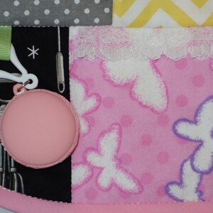 May include: A pink and white baby blanket with a variety of patterns, including polka dots, zigzags, and butterflies. The blanket has a white lace trim and a pink and white tag with a small pink circle attached.