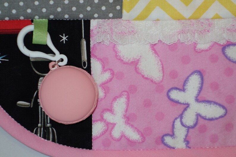 May include: A pink and white baby blanket with a variety of patterns, including polka dots, zigzags, and butterflies. The blanket has a white lace trim and a pink and white tag with a small pink circle attached.