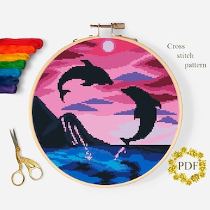 May include: Cross-stitch pattern featuring two leaping dolphins against a pink and purple sunset sky. The design is framed in a wooden hoop, with the text "Cross stitch pattern" and a "PDF" logo. Colorful embroidery floss and gold scissors are also visible.