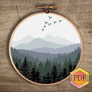 Peut inclure: Un motif de point de croix repr&eacute;sentant un paysage montagneux brumeux avec une for&ecirc;t au premier plan et un vol d'oiseaux au-dessus. Le motif est dans des tons de bleu, vert et noir.