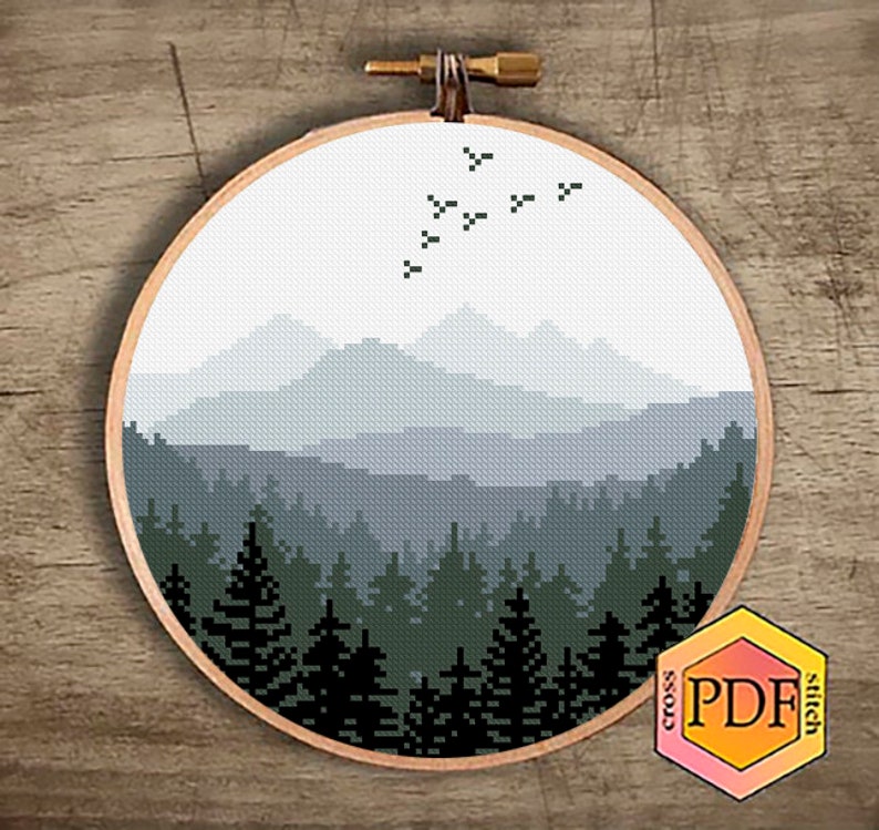 Peut inclure: Un motif de point de croix repr&eacute;sentant un paysage montagneux brumeux avec une for&ecirc;t au premier plan et un vol d'oiseaux au-dessus. Le motif est dans des tons de bleu, vert et noir.