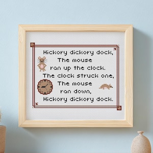 May include: A framed cross-stitch featuring the nursery rhyme "Hickory Dickory Dock." The text is in dark brown, with illustrations of a mouse and a clock. The frame is light wood, and the background is a light blue wall.