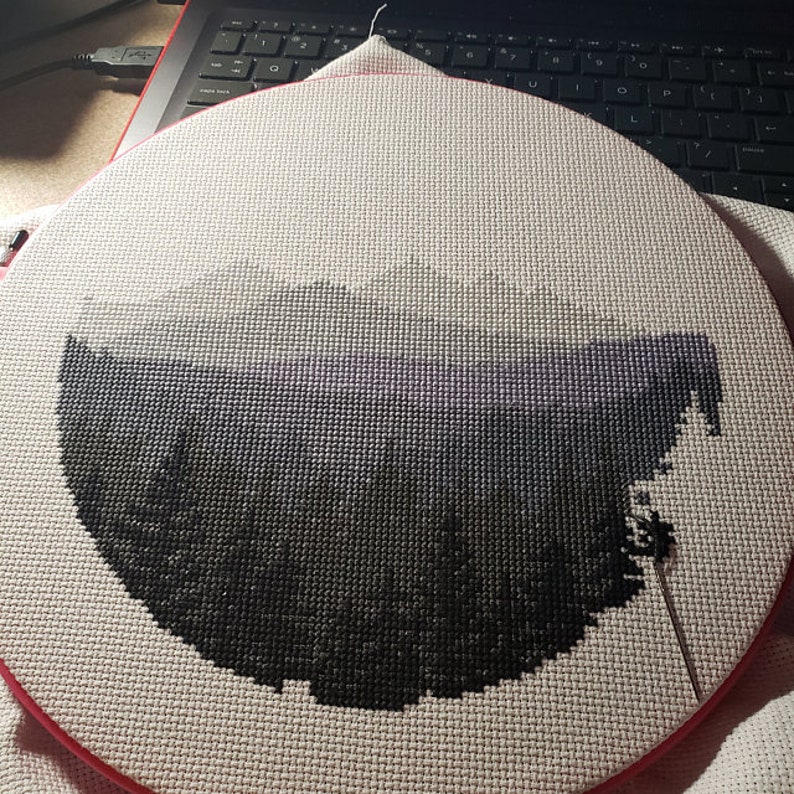Peut inclure: Gros plan d'un projet de point de croix partiellement termin&eacute; repr&eacute;sentant un paysage d&eacute;grad&eacute; en noir et blanc. Le projet est mont&eacute; dans un cercle de broderie rond avec un anneau ext&eacute;rieur rouge.