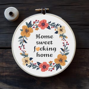 Könnte beinhalten: Ein Kreuzstich-Stickrahmen mit dem Text "Home sweet f*cking home", umgeben von einem floralen Kranz in Orange, Gelb und Grün. Eine Spule weißen Fadens liegt daneben.