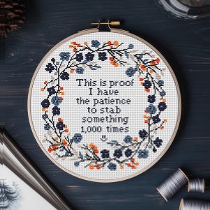 Peut inclure: Un cercle de broderie au point de croix avec une couronne florale bleue, orange et blanche. Le texte dit : « C'est la preuve que j'ai la patience de poignarder quelque chose 1 000 fois ». Une pièce décorative et humoristique.