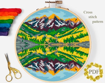 May include: Cross stitch pattern of a mountain landscape with a lake reflecting the mountains. The pattern is in a hoop and includes a variety of colors, including green, yellow, blue, and brown.
