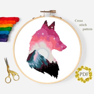 Puede incluir: Patr&oacute;n de punto de cruz de la silueta de la cabeza de un lobo con una monta&ntilde;a y una luna en su interior. El lobo es rosa, morado y azul con estrellas blancas. La monta&ntilde;a es azul y verde. La luna es blanca. El patr&oacute;n est&aacute; en un aro de bordado blanco. El texto "Cross stitch pattern" y "PDF" es visible en la imagen.