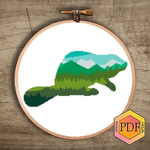 May include: Cross stitch pattern featuring a beaver silhouette filled with a green, blue, and white landscape design. The pattern is printed on a white background and framed in a wooden embroidery hoop.