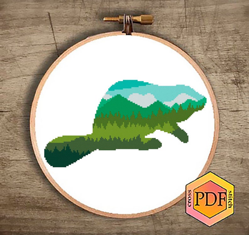 May include: Cross stitch pattern featuring a beaver silhouette filled with a green, blue, and white landscape design. The pattern is printed on a white background and framed in a wooden embroidery hoop.
