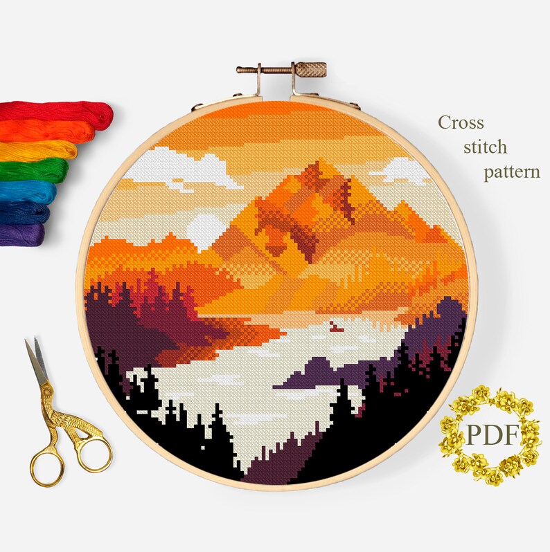 Op de afbeelding: Kruissteekpatroon van een berglandschap met een zonsondergangshemel. De bergen zijn oranje en geel, met donkergroene bomen op de voorgrond. De lucht is een gradi&euml;nt van oranje, geel en wit.