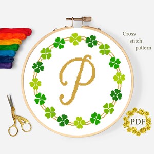 May include: Cross stitch pattern featuring a gold letter P surrounded by a wreath of green four-leaf clovers. The pattern is displayed in a wooden embroidery hoop.