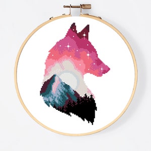 Puede incluir: Patr&oacute;n de punto de cruz de una silueta de lobo con una monta&ntilde;a y un cielo estrellado en su interior. El lobo es rosa, morado y azul, con un contorno negro. El patr&oacute;n est&aacute; en un aro de bordado de madera.