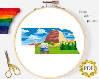 May include: A cross-stitch pattern of a landscape scene, featuring a blue sky with white clouds, a brown mountain, and a green field. The design is in a wooden embroidery hoop. The text "Cross stitch pattern" is visible.