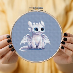 Op de afbeelding: Een borduurring met kruissteek met een witte draak met grote blauwe ogen en vleugels, tegen een lichtblauwe achtergrond. De draak staat in het midden van de ring, die wordt vastgehouden door handen met zwarte nagellak.