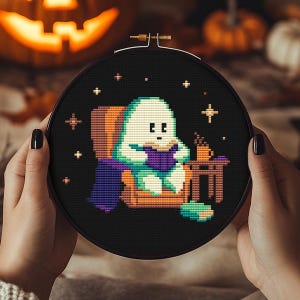 May include: A black cross-stitch embroidery hoop featuring a pixelated ghost reading a book while seated in a chair. The ghost is wearing a purple cape and is holding a book. A small table with a cup of coffee sits beside the ghost. The background is black with star accents.