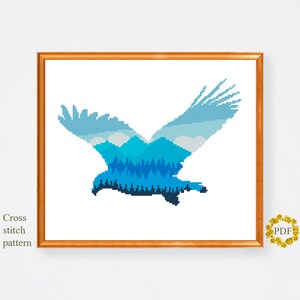 Pu&ograve; includere: Schema di punto croce di un'aquila blu con un paesaggio di montagna e foresta all'interno della sua silhouette.