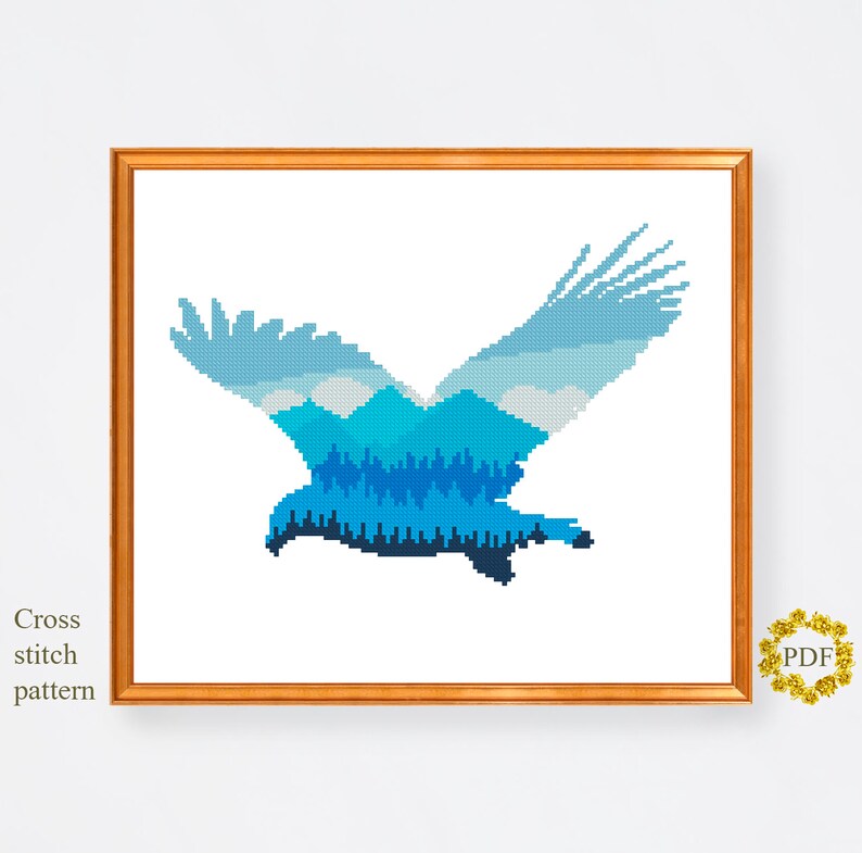 Pu&ograve; includere: Schema di punto croce di un'aquila blu con un paesaggio di montagna e foresta all'interno della sua silhouette.