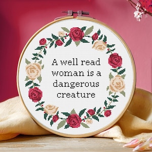 May include: Cross-stitch embroidery in a wooden hoop. The design features the quote "A well read woman is a dangerous creature" surrounded by a wreath of red and cream-colored roses and green leaves. The background is a soft pink.