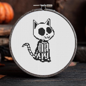 Pode incluir: Ponto cruz preto e branco de um esqueleto de gato num bastidor de bordar de madeira. O gato tem grandes órbitas redondas e uma caixa torácica detalhada. Uma pequena abóbora laranja é visível no fundo, sugerindo um tema de Halloween.