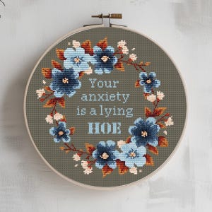 Może przedstawiać: Obręcz do haftu krzyżykowego z kwiatowym wieńcem z niebieskich kwiatów i brązowych liści. Tekst "Your anxiety is a lying HOE" jest wyhaftowany pośrodku na szarym tle.