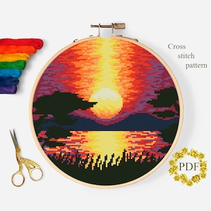 May include: Cross stitch pattern of a sunset over a lake with trees in the foreground. The sunset is a vibrant orange, yellow, and red. The lake is a dark blue with reflections of the sunset. The trees are a dark green. The pattern is in a round embroidery hoop.