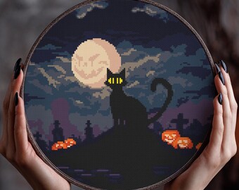 May include: A cross-stitch embroidery hoop featuring a Halloween scene. A black cat with yellow eyes sits atop a hill, silhouetted against a full moon. The background depicts a night sky with clouds and pumpkins.