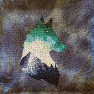 May include: Cross-stitch embroidery of a wolf's head with a mountain range and starry sky inside the silhouette. The wolf is outlined in dark blue and the mountain range is in shades of blue and green.