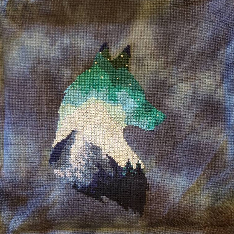 May include: Cross-stitch embroidery of a wolf's head with a mountain range and starry sky inside the silhouette. The wolf is outlined in dark blue and the mountain range is in shades of blue and green.