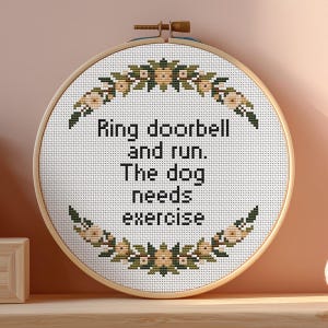 May include: A round, cross-stitch embroidery hoop with the text "Ring doorbell and run. The dog needs exercise." The design features a floral border in shades of green, brown, and cream. The hoop is made of wood and has a brass-colored clasp.