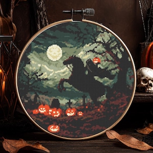 May include: A cross-stitch embroidery in a wooden hoop shows a Halloween scene. A black horse rears under a full moon, with carved pumpkins in the foreground. The background features a dark forest with a skull and pumpkins.