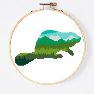 May include: Cross stitch pattern of a beaver silhouette filled with a green forest and blue sky landscape. The pattern is displayed in a wooden embroidery hoop. The text "Cross stitch PDF" is below the hoop.