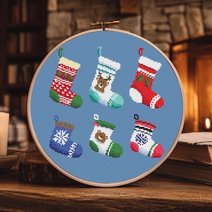 May include: A cross-stitch embroidery hoop featuring six miniature Christmas stockings. Each stocking is a different color and design, including snowflakes, reindeer, and geometric patterns. The hoop is set against a blue background.