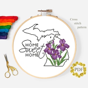Może przedstawiać: Wzór haftu krzyżykowego w drewnianym tamborku przedstawia zarys stanu Michigan, słowa „Home Sweet Home” i fioletowe kwiaty irysów. Widoczne są nici do haftu i złote nożyczki.