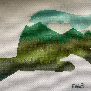 May include: Cross-stitch embroidery of a beaver silhouette with a green, blue, and white mountain landscape inside. The design is on white fabric and signed "Feb'21".