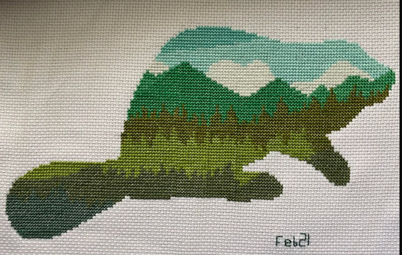 May include: Cross-stitch embroidery of a beaver silhouette with a green, blue, and white mountain landscape inside. The design is on white fabric and signed "Feb'21".
