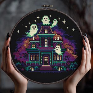 以下が含まれることがあります： お化け屋敷のシーンを描いたクロスステッチ刺繍枠。家は紫とティールで、星が輝く黒い夜空を背景にしています。デザインは木製の刺繍枠に収められています。