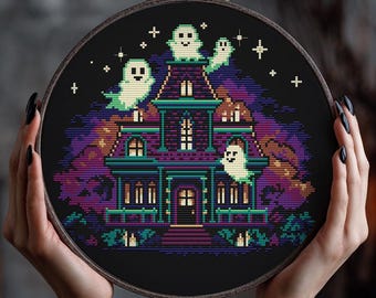 May include: A cross-stitch embroidery hoop featuring a spooky haunted house scene with pixelated ghosts. The house is purple and teal, set against a black night sky with stars. The design is held in a wooden embroidery hoop.