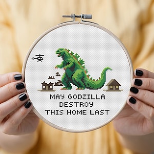 Puede incluir: Arte de bordado en punto de cruz en un aro que muestra a Godzilla verde destruyendo edificios. El texto dice: "MAY GODZILLA DESTROY THIS HOME LAST". El diseño también incluye un helicóptero. La obra de arte se muestra en un aro de madera.
