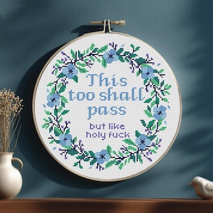 Puede incluir: Bordado de punto de cruz en un aro de madera. El diseño presenta el texto "This too shall pass but like holy fuck" en azul, rodeado por una corona floral de flores azules y hojas verdes. El fondo es blanco.