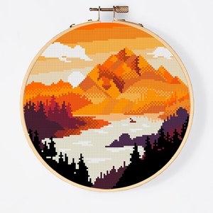 Op de afbeelding: Kruissteekpatroon van een berglandschap met een zonsondergangshemel. De bergen zijn oranje en bruin, met een meer op de voorgrond. De lucht is oranje en geel, met witte wolken. Het patroon is in een ronde borduurring.